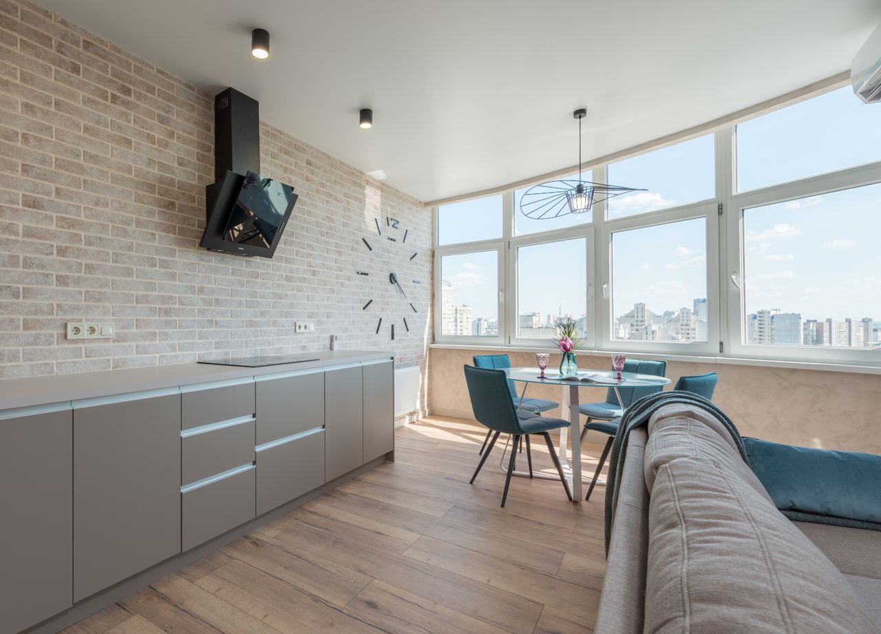 Interior design of new stylish studio consisting of spacious kitchen area near comfortable couch and dining zone with clock on brick wall and big windows